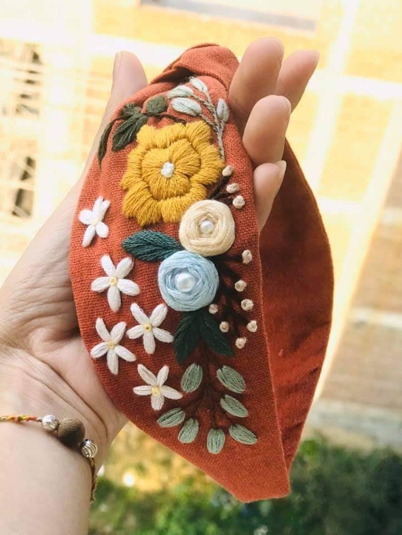 May include: A rust-colored fabric headband with a knotted design, featuring embroidered flowers in shades of yellow, white, blue, and green.