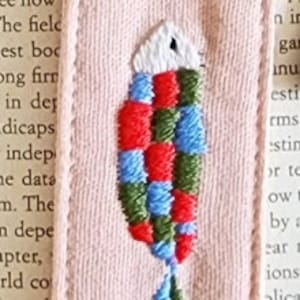 May include: A pink fabric bookmark with a colorful embroidered fish design. The fish is red, green, blue, and white. The bookmark has a loop at the top for attaching to a book.
