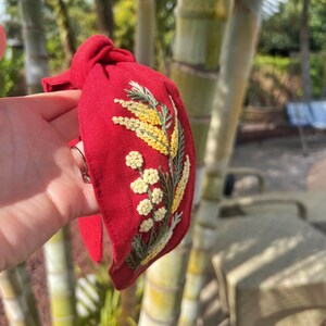 May include: Red fabric headband with a knotted bow and embroidered yellow flowers.