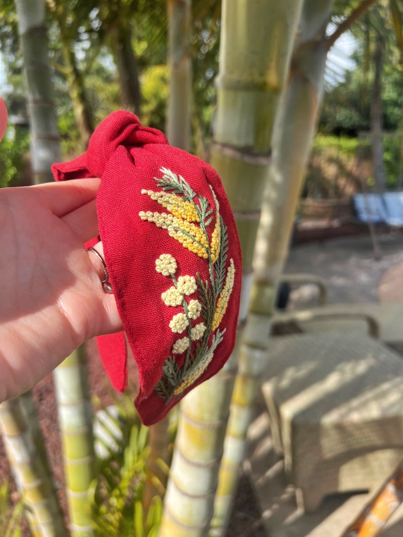 May include: Red fabric headband with a knotted bow and embroidered yellow flowers.