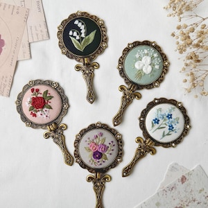May include: Five ornate, hand-held mirrors with embroidered floral designs. Each mirror has a gold-toned frame and handle. The floral designs include lilies of the valley, white roses, forget-me-nots, and a bouquet of pink and purple roses.