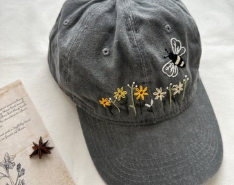 Può includere: Un cappello da baseball grigio con fiori gialli ricamati e un'ape. Il cappello ha una visiera curva e un aspetto leggermente usurato.