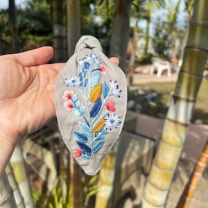 May include: A gray fabric headband with a knotted top and an embroidered floral design in shades of blue, yellow, white, and pink.