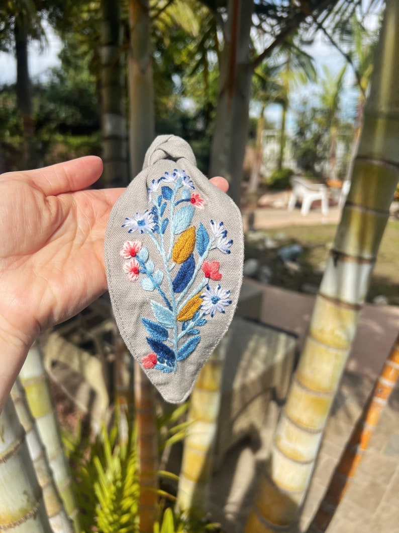 May include: A gray fabric headband with a knotted top and an embroidered floral design in shades of blue, yellow, white, and pink.