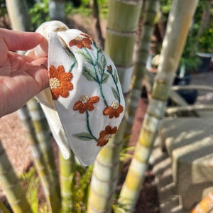 May include: White fabric headband with a knotted design and embroidered orange flowers and green leaves.