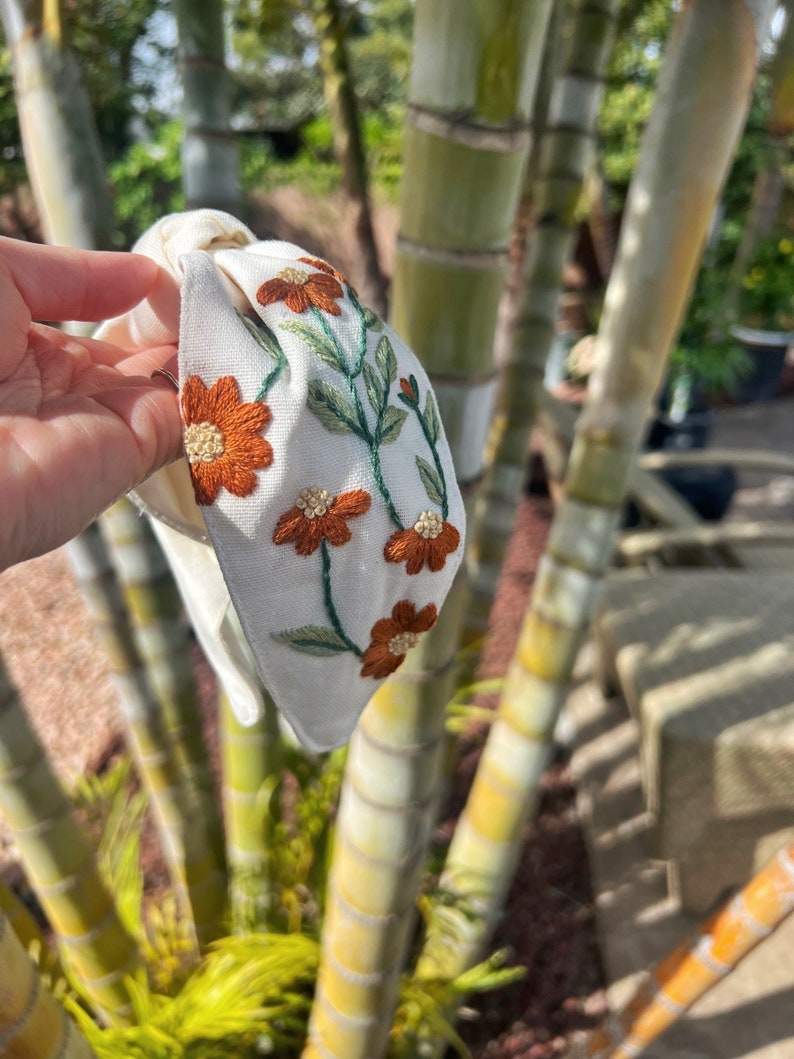 May include: White fabric headband with a knotted design and embroidered orange flowers and green leaves.