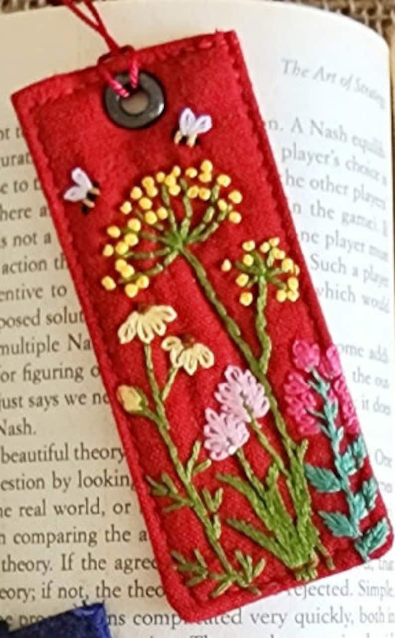 May include: Red felt bookmark with embroidered wildflowers in shades of yellow, pink, green, and white. The bookmark has a metal ring at the top.