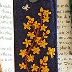 May include: A dark blue fabric bookmark with a yellow embroidered flower design and a small bee. The bookmark has a brown leather strap and a metal ring.