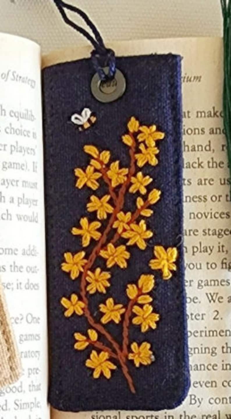 May include: A dark blue fabric bookmark with a yellow embroidered flower design and a small bee. The bookmark has a brown leather strap and a metal ring.