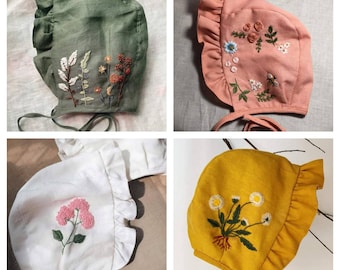 May include: Four different styles of handmade baby bonnets with embroidered floral designs. The bonnets are made of a soft, natural fabric and come in green, pink, white, and yellow.