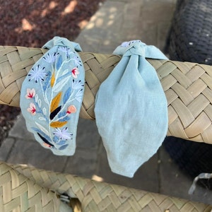 May include: Two light blue fabric headbands. One headband has a floral embroidery design in shades of blue, pink, yellow, and white. The other headband is plain.