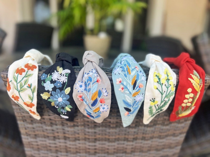 May include: Six fabric headbands with knotted ends, each featuring a different floral embroidery design in various colors, including white, black, gray, blue, yellow, and red.