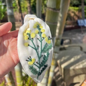 May include: White fabric headband with a hand-embroidered floral design featuring yellow flowers and green leaves.