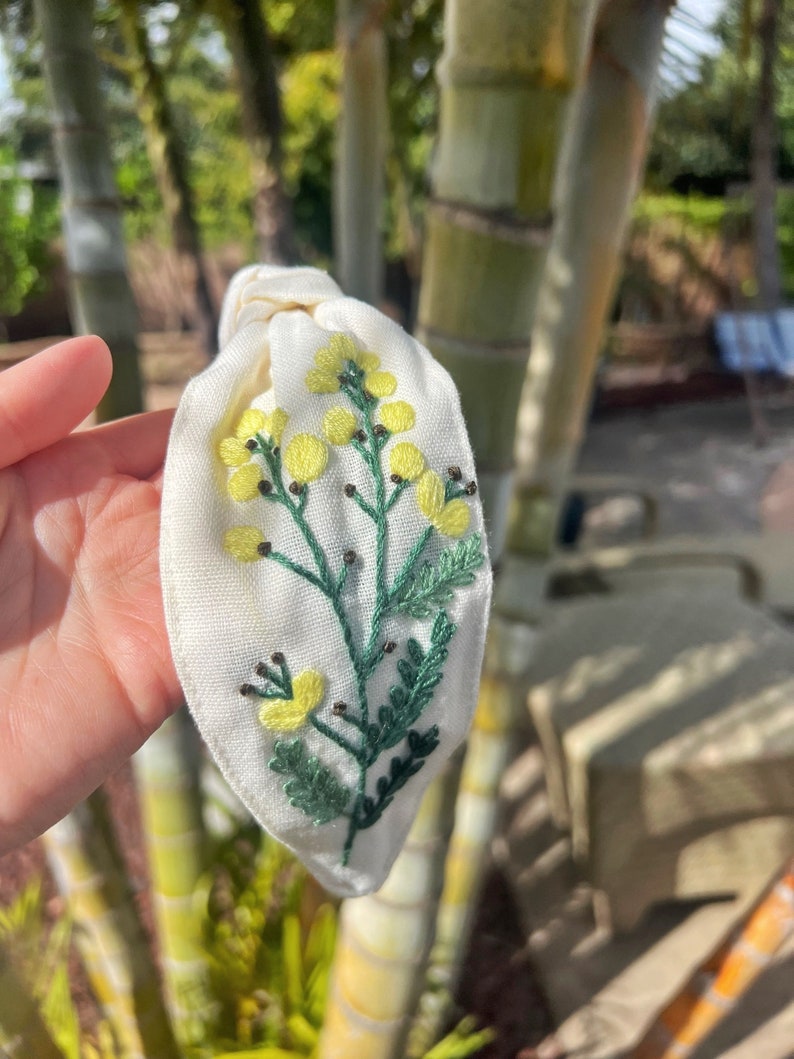 May include: White fabric headband with a hand-embroidered floral design featuring yellow flowers and green leaves.