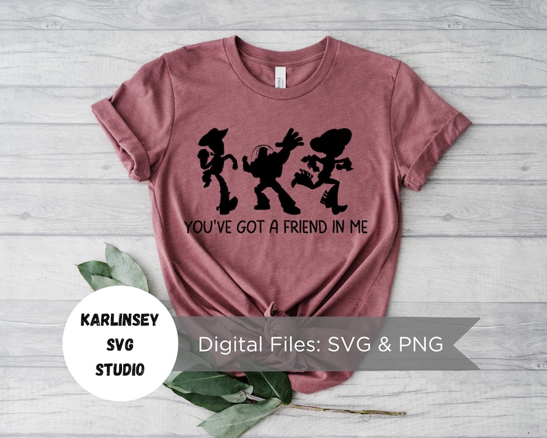 Op de afbeelding: Een maronkleurig T-shirt met een zwart silhouet van de Toy Story-personages Woody, Buzz Lightyear en Jessie die dansen. De tekst "You've got a friend in me" is onder de personages gedrukt. De tekst "KARLINSEY SVG STUDIO" is gedrukt in een cirkel linksonder in de afbeelding. De tekst "Digital Files: SVG & PNG" is gedrukt in een wit rechthoek rechtsonder in de afbeelding.