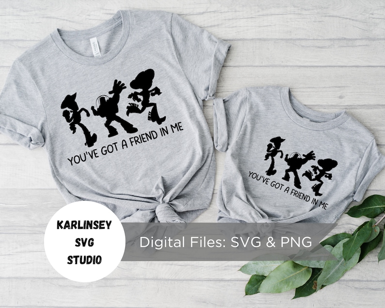 Op de afbeelding: Twee grijze T-shirts met een zwart silhouet van de Toy Story-personages Woody, Buzz Lightyear en Jessie die dansen. De tekst "You've got a friend in me" staat op de T-shirts gedrukt.