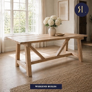 May include: A light-colored wooden dining table with a trestle base. The table is rectangular and has a natural wood finish. A vase of white flowers sits on the table. The text "WEEKEND BUILDS" is at the bottom.