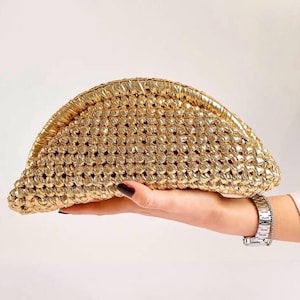 May include: A gold clutch bag with a woven pattern. The bag is held in a hand, displaying its crescent shape and textured surface. The metallic sheen of the bag suggests it is a formal or evening accessory.