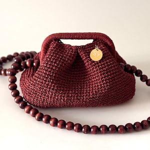 May include: A burgundy crocheted handbag with a matching handle and a beaded wooden strap. The bag has a small gold-colored charm attached. The beads are a dark brown color. The bag is on a white background.