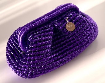 May include: A vibrant purple clutch bag with a woven texture. The bag features a rounded shape with a fold-over flap. A small, circular gold-toned charm with the letters "SY" hangs from the side. The bag is made of a shiny material.