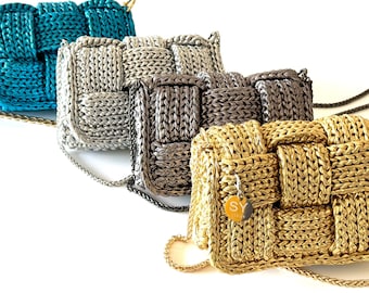 May include: Four woven-style crossbody bags in teal, silver, charcoal, and gold. Each bag features a chain strap and a rectangular shape with a woven pattern. The gold bag has a small charm with the letters "SY".
