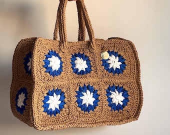 May include: A brown crocheted tote bag with blue and white flower designs. The bag has two handles and a small brown tag with the letters "SY" on it.