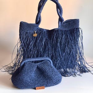 May include: A set of two blue crocheted bags. The larger bag has a fringe trim and a top handle. The smaller bag has a top handle and a label that reads "SY DESIGN".