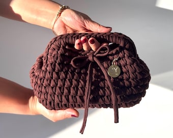 Può includere: Una pochette marrone scuro, realizzata a mano all'uncinetto. La borsa presenta una forma arrotondata, un manico annodato, una chiusura con coulisse e un piccolo ciondolo circolare con le iniziali "SY".