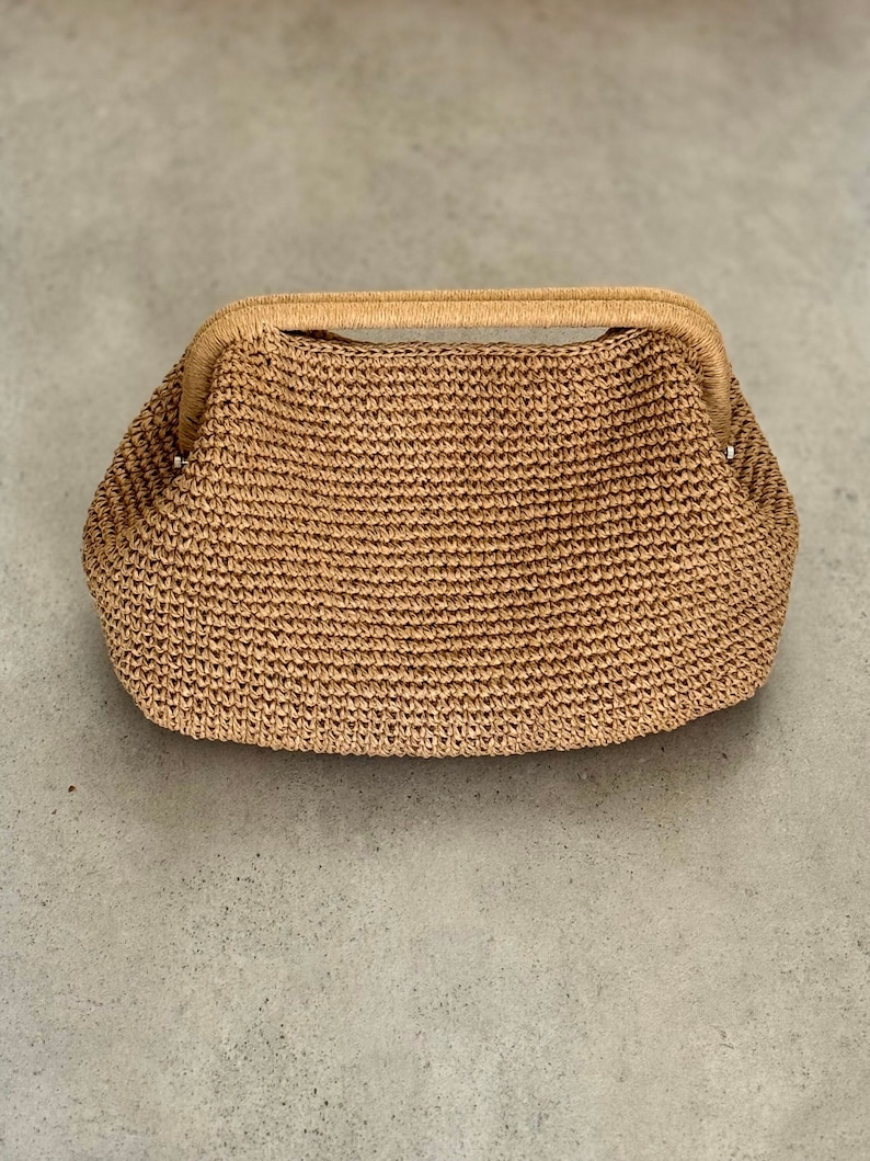 May include: A woven straw handbag with a curved handle. The bag is a light tan color and has a textured surface. The handle is a lighter shade of tan and is also woven. The bag is sitting on a gray surface.