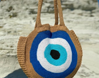 May include: A crocheted tote bag with a large blue evil eye design. The bag is brown and has a white, blue, and turquoise eye with a black pupil.