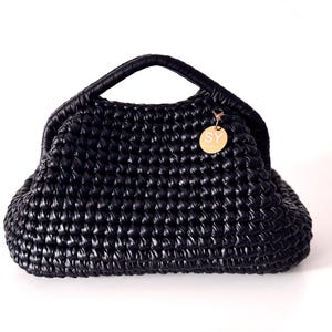 Peut inclure: Sac à main noir tissé, de forme arrondie, avec une poignée supérieure. Un petit charme doré avec les lettres "SY" est suspendu sur le côté. Le sac a un aspect texturé et brillant, et une conception structurée.