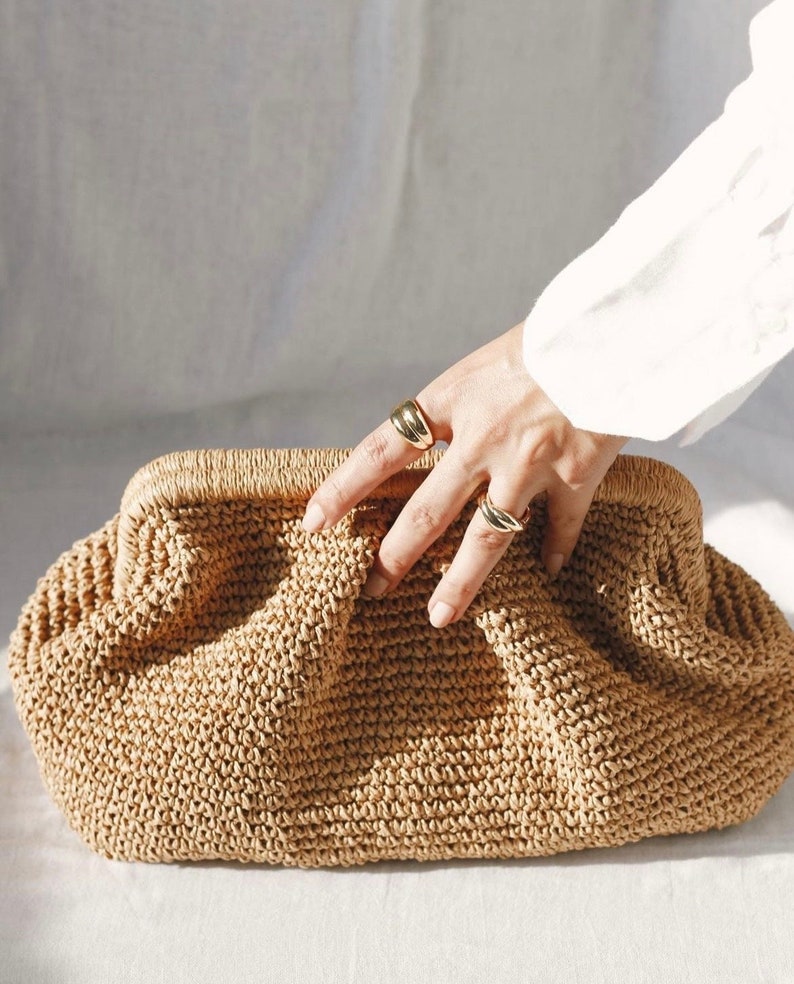 Pu&ograve; includere: Una pochette in vimini di colore marrone chiaro con un design plissettato e gonfio. La borsa &egrave; realizzata in un materiale intrecciato fitto e ha una superficie liscia leggermente testurizzata.