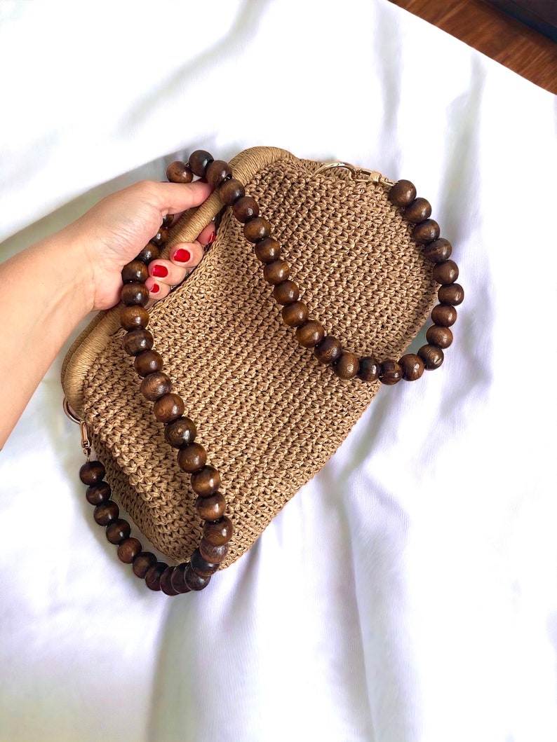 May include: A woven straw handbag with a wooden beaded handle. The bag is a light brown color with a matching woven texture. The handle is made of large, dark brown wooden beads.