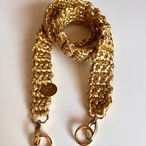 May include: Gold-colored crocheted bag strap with two gold-colored metal clasps. The strap has a small gold-colored tag with the text "SY Studio" on it.