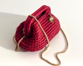 May include: A red woven handbag with a gold chain strap. The small purse has a rounded shape with a handle at the top. A gold-colored charm with the letters "SY" hangs from the side.