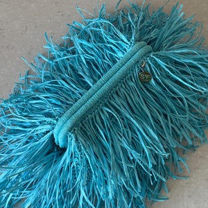 May include: A bright turquoise clutch bag featuring a dense, fringed exterior made of thin, ribbon-like strands. The bag has a structured, rounded top closure with a central seam and a small, circular gold-toned metal charm engraved with the letters SY.