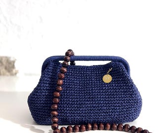 May include: A navy blue crocheted handbag with a wooden bead strap. The bag has a rounded shape and a small gold-colored charm. The wooden beads are dark brown. The bag is sitting on a white surface.
