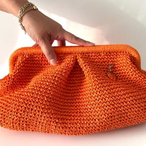 May include: Bright orange woven clutch with a rounded shape and a clasp closure. The bag features a textured, open weave design and a small charm. A stylish accessory for carrying essentials.