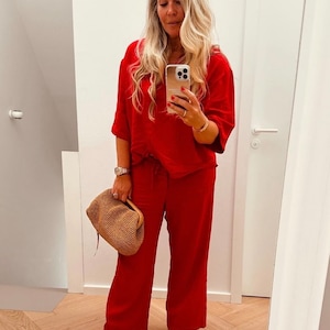 May include: A woman in a mirror selfie wearing a red outfit, including a short-sleeved top and wide-leg pants. She holds a woven handbag and wears brown sandals. Her long blonde hair is styled down.