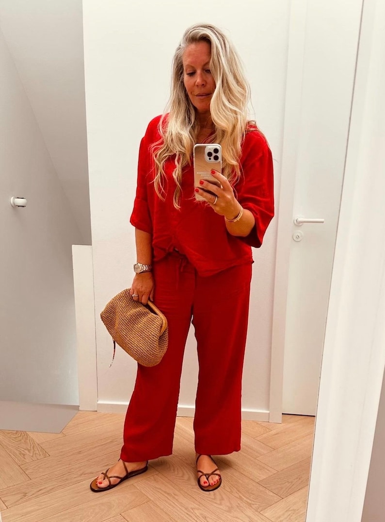 May include: A woman in a mirror selfie wearing a red outfit, including a short-sleeved top and wide-leg pants. She holds a woven handbag and wears brown sandals. Her long blonde hair is styled down.