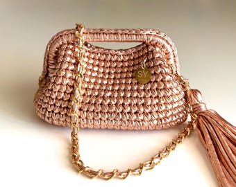 May include: A rose gold crocheted handbag with a gold chain strap and a matching tassel. The bag has a structured shape with a handle and a small circular tag with the letters "SY".