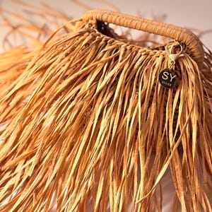 May include: A handcrafted, light brown woven bag with a sturdy handle. The bag features long, fringed edges and a small, circular tag with the letters "SY DESIGN". The bag is made of natural fibers.
