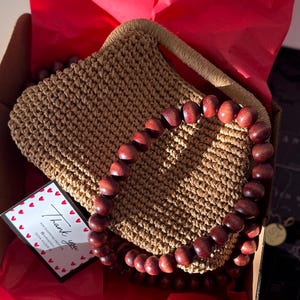 May include: A handcrafted, light brown crochet handbag with a matching handle and a beaded wooden accent. The bag is presented in a cardboard box with red tissue paper and a thank you card.