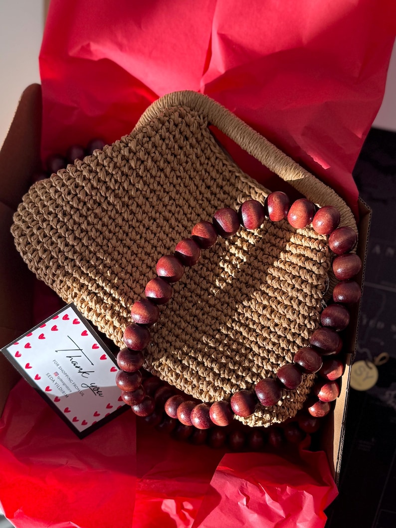 May include: A handcrafted, light brown crochet handbag with a matching handle and a beaded wooden accent. The bag is presented in a cardboard box with red tissue paper and a thank you card.