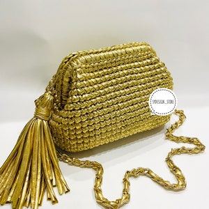 Può includere: Una pochette dorata intrecciata con una tracolla a catena dorata e un pendente a nappa dorato. La borsa ha un design intrecciato testurizzato e una forma rettangolare.