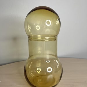 May include: A translucent amber-colored glass vessel consisting of a rounded base with a narrower neck and a large, spherical glass lid that sits securely on top, creating a smooth, continuous silhouette with a warm, golden-brown hue.