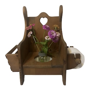 May include: A brown wooden toilet paper holder featuring a high backrest with a heart-shaped cutout. The unit includes a side storage bin, a central circular opening holding a glass jar with pink and white zinnia flowers, and a side roll holder.