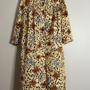 May include: A knee-length housecoat featuring a yellow base with a repeating pattern of brown floral motifs and black speckled accents. The garment has three-quarter length sleeves, a collared neckline, and a full-length front button closure.