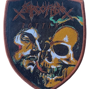 May include: A black and brown embroidered patch with the band name "CARCASS" and the word "ROTTING" in white lettering. The patch features a skull and a human face with a red dot on the cheek.