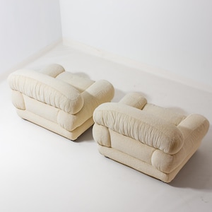 May include: Two cream-coloured, upholstered ottomans. Each ottoman has a rectangular base with rounded corners and a series of rounded, cushioned sections. The fabric has a textured appearance, and the ottomans are placed on a white surface.